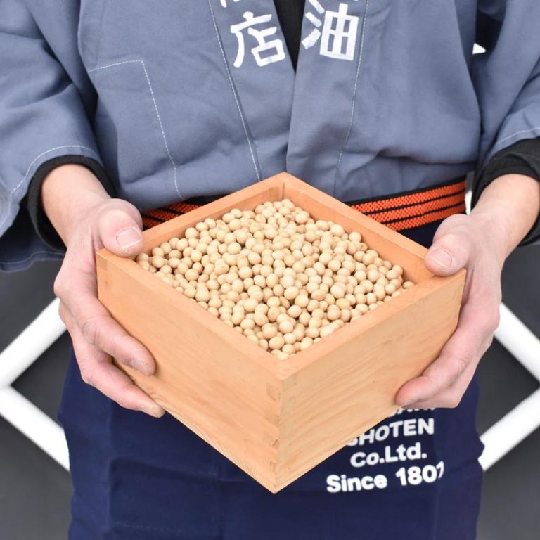 Una persona in abito tradizionale da lavoro giapponese (con grembiule blu e fasce rosse) regge una cassa di legno colma di fagioli di soia gialli, ingredienti fondamentali per la produzione di salsa di soia e miso. La cassa, in legno naturale, porta il marchio ‘Yagisawa Shoten Co. Ltd. Since 1807’, a testimoniare la lunga storia dell’azienda. I fagioli, lucidi e uniformi, rappresentano l’inizio di un processo di fermentazione che trasformerà materie prime semplici in prodotti ricchi di umami e tradizione.