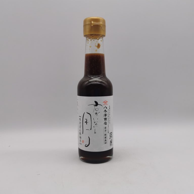 Bottiglia in vetro scuro di salsa ponzu allo yuzu Kimi Ga Inai To Komaru, prodotta da Yagisawa Shoten, con tappo dorato e etichetta bianca decorata da calligrafia giapponese a mano in nero. L’etichetta riporta il nome del prodotto in caratteri tradizionali e il logo dell’azienda, simbolo di una storia di resilienza e rinascita dopo il terremoto del 2011. La bottiglia, fotografata su sfondo neutro, evoca l’eleganza artigianale dei condimenti giapponesi, con il liquido scuro che traspare attraverso il vetro.
