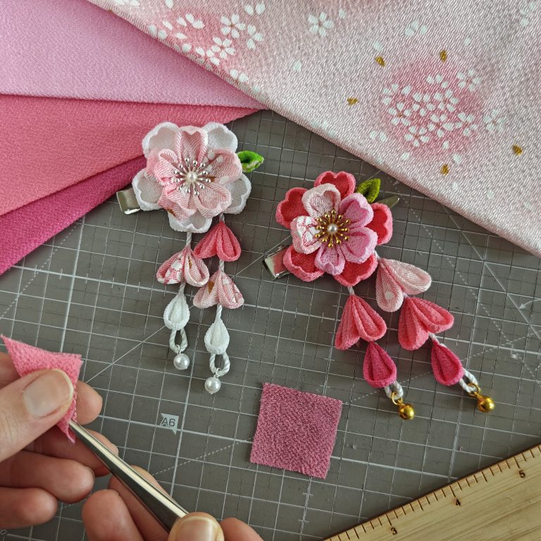 Due fermagli tsumami zaiku a forma di fiori di ciliegio (sakura) in fase di realizzazione su un tavolo da lavoro. Il fermaglio a sinistra è in toni rosa chiaro e bianco, con petali pendenti e perle decorative, mentre quello a destra è rosa intenso con campanelle dorate. Una mano tiene un pezzo di stoffa rosa e una pinza, pronta per tagliare il tessuto su un tappetino da taglio con griglia di misurazione.