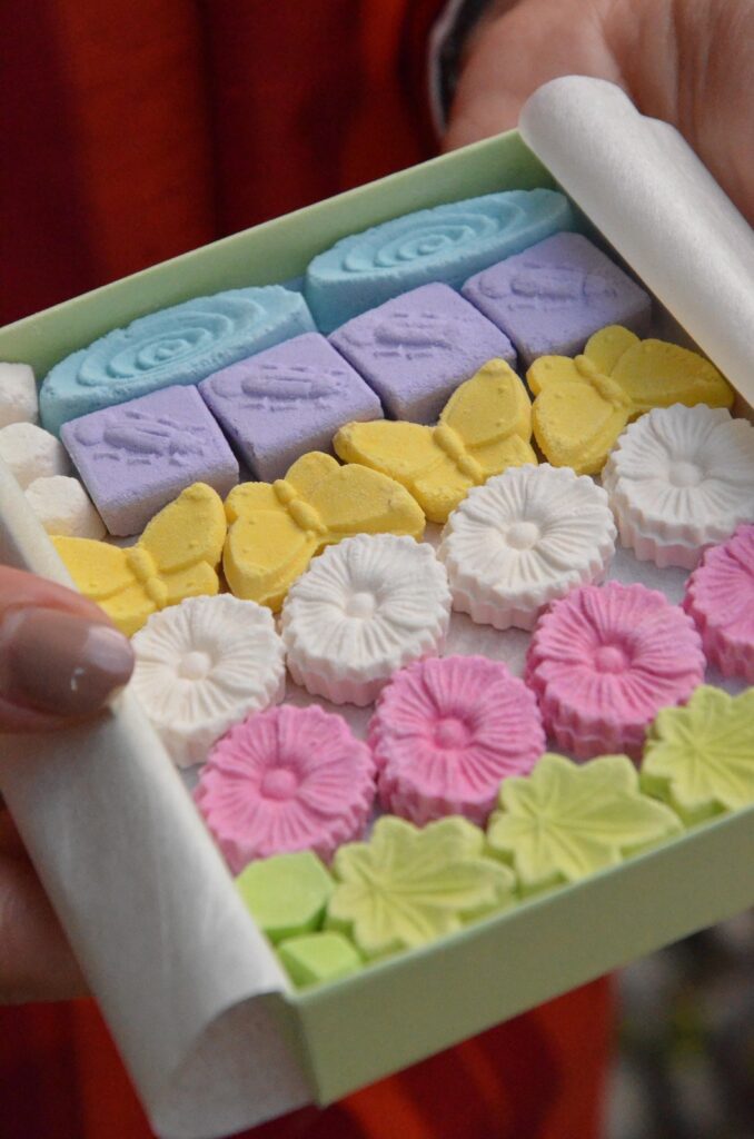 Una scatola verde piena di biscotti decorati a forma di fiori e farfalle di vari colori, tenuta da una mano con smalto rosa sulle unghie.