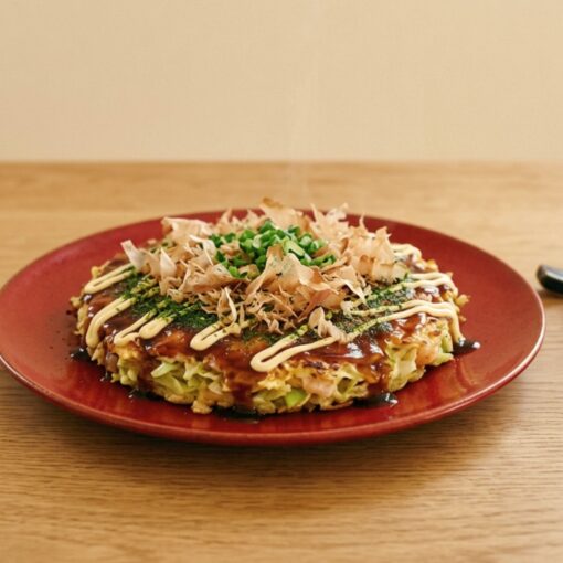 Okonomiyaki su piatto rosso con salsa Otafuku, maionese Kewpie, katsuobushi e cipollotto, servito con bacchette.