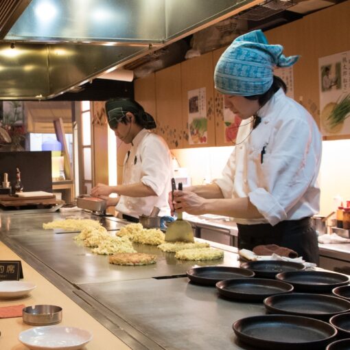 Due chef preparano okonomiyaki su una piastra calda, uno indossa un cappello blu e l'altro un cappello verde, in una cucina giapponese.
