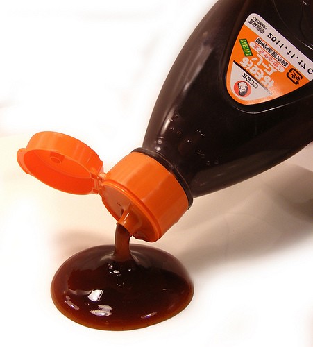 Una bottiglia di salsa okonomiyaki Otafuku con il tappo arancione aperto, mentre la salsa scura e densa cola su una superficie bianca, creando una pozza lucida.
