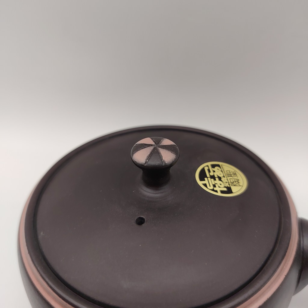 Tappo di una kyusu in ceramica nera con dettagli rosa, un piccolo foro per il vapore e un'etichetta dorata con caratteri giapponesi.