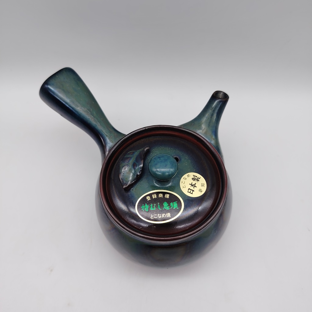 Kyusu in ceramica verde lucida con manico laterale e beccuccio, decorato con una piccola fogliolina in rilievo sul coperchio. Etichetta giapponese sul coperchio indica "Made in Japan" e "Tokoname-yaki".
