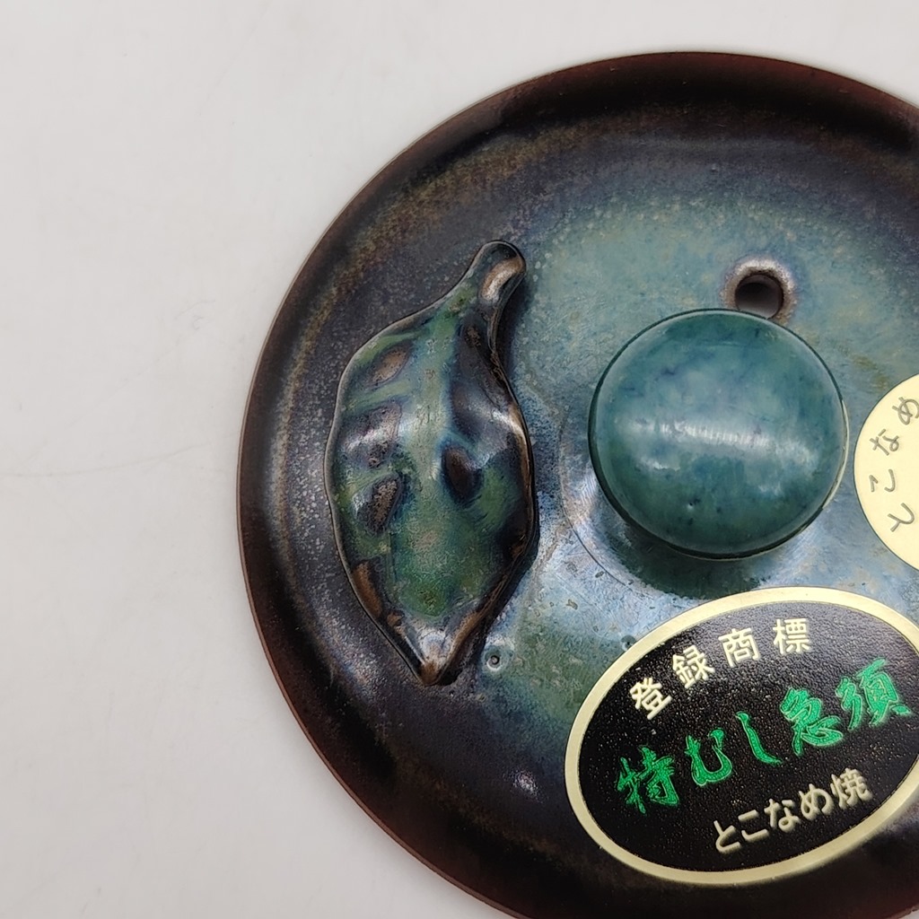 Tappo di kyusu in ceramica verde con decorazione a foglia e pomello rotondo, etichetta con testo giapponese e "tokoname-yaki".