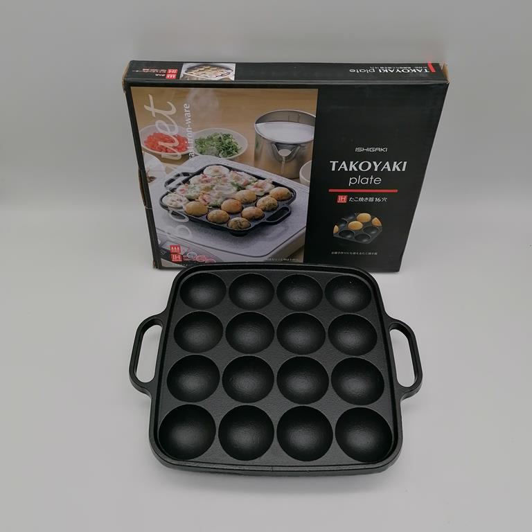 Padella per takoyaki in ghisa Ishigaki con 16 fori, nera, con manici, su sfondo bianco. La confezione è visibile sullo sfondo.