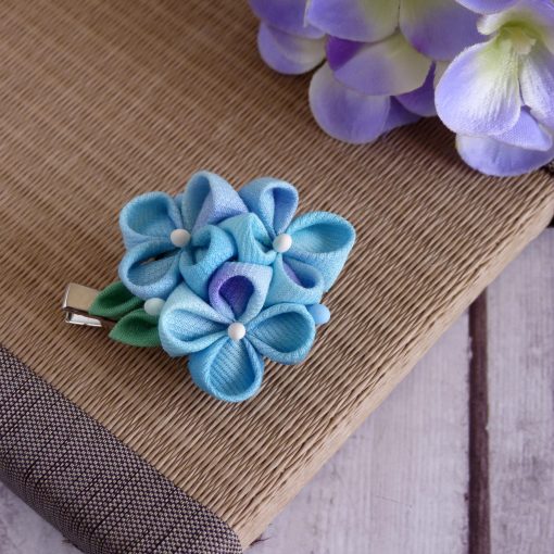 Spilla floreale Tsumami Zaiku azzurra e viola, con dettagli verdi, su sfondo di paglia e fiori di glicine viola, ideale per laboratori estivi di Momo Kanzashi.