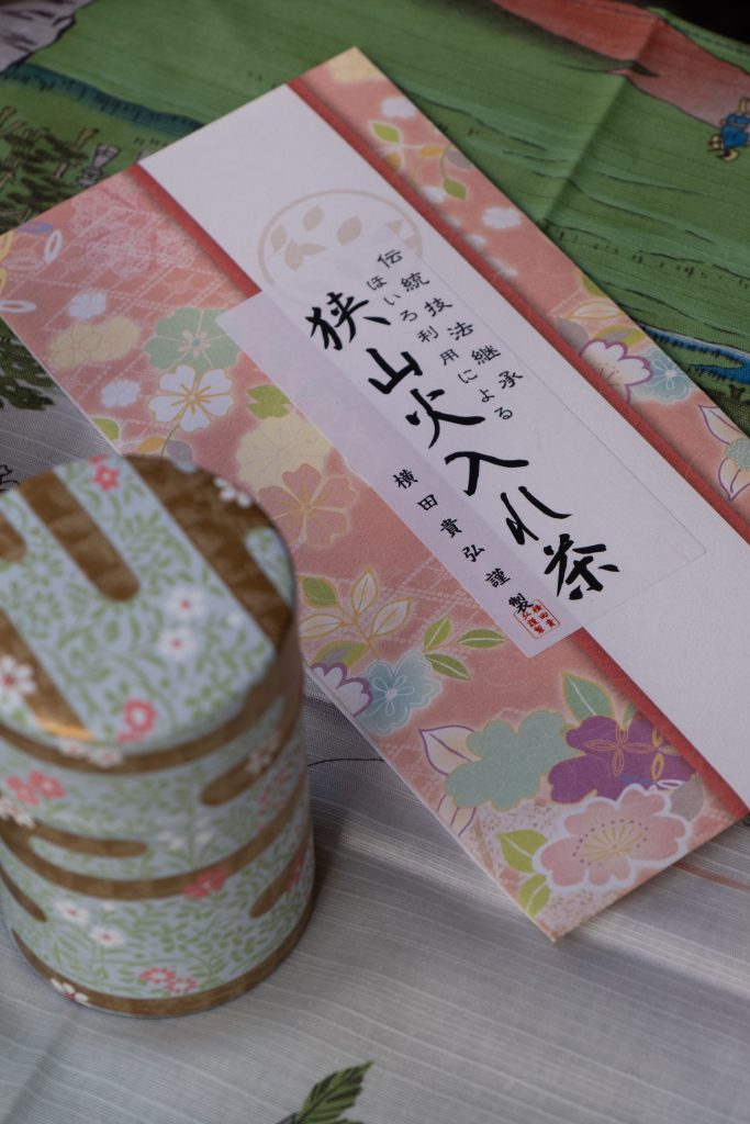 Un regalo giapponese tradizionale con confezione regalo di carta decorata con motivi floreali e un barattolo di latta a tre strati con coperchio dorato, posto su una superficie con un tessuto a righe.