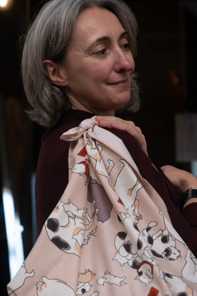 Donna con capelli grigi corti sorride con un foulard rosa con stampa di gatti legato a tracolla, con un maglione bordeaux.