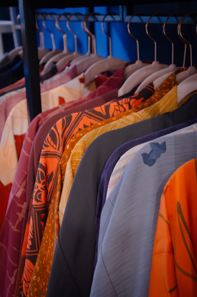 Colorati kimono giapponesi appesi a un bastone, con motivi floreali e geometrici in tonalità vivaci come arancione, blu e viola, disposti su una mensola blu scuro.