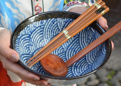 A person in a colorful robe decorated with dragons and waves holds a blue bowl with a sea wave pattern, with wooden chopsticks and a wooden spoon resting on top.