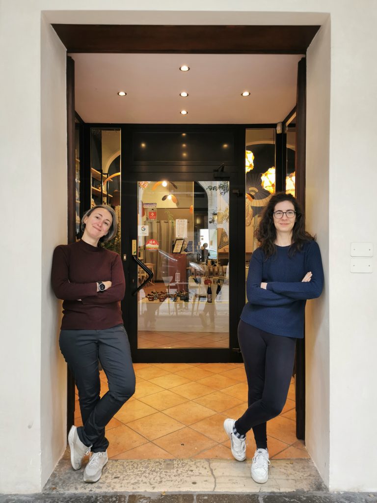 Due donne sorridenti con braccia incrociate, una con maglione marrone e l'altra con maglione blu, in piedi accanto a una porta aperta di un negozio con una vetrina che mostra l'interno.
