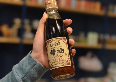 A hand holding a bottle of soy sauce with a Japanese label, with blurred background shelves.