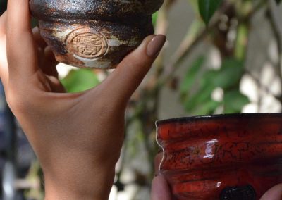 Two hands holding two small handcrafted ceramic cups, one dark with engravings and one bright red, with green leaves in the background.