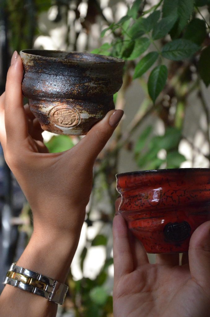 Due mani tengono due piccole tazze di ceramica artigianale, una scura con incisioni e una rossa vivace, con foglie verdi sullo sfondo.